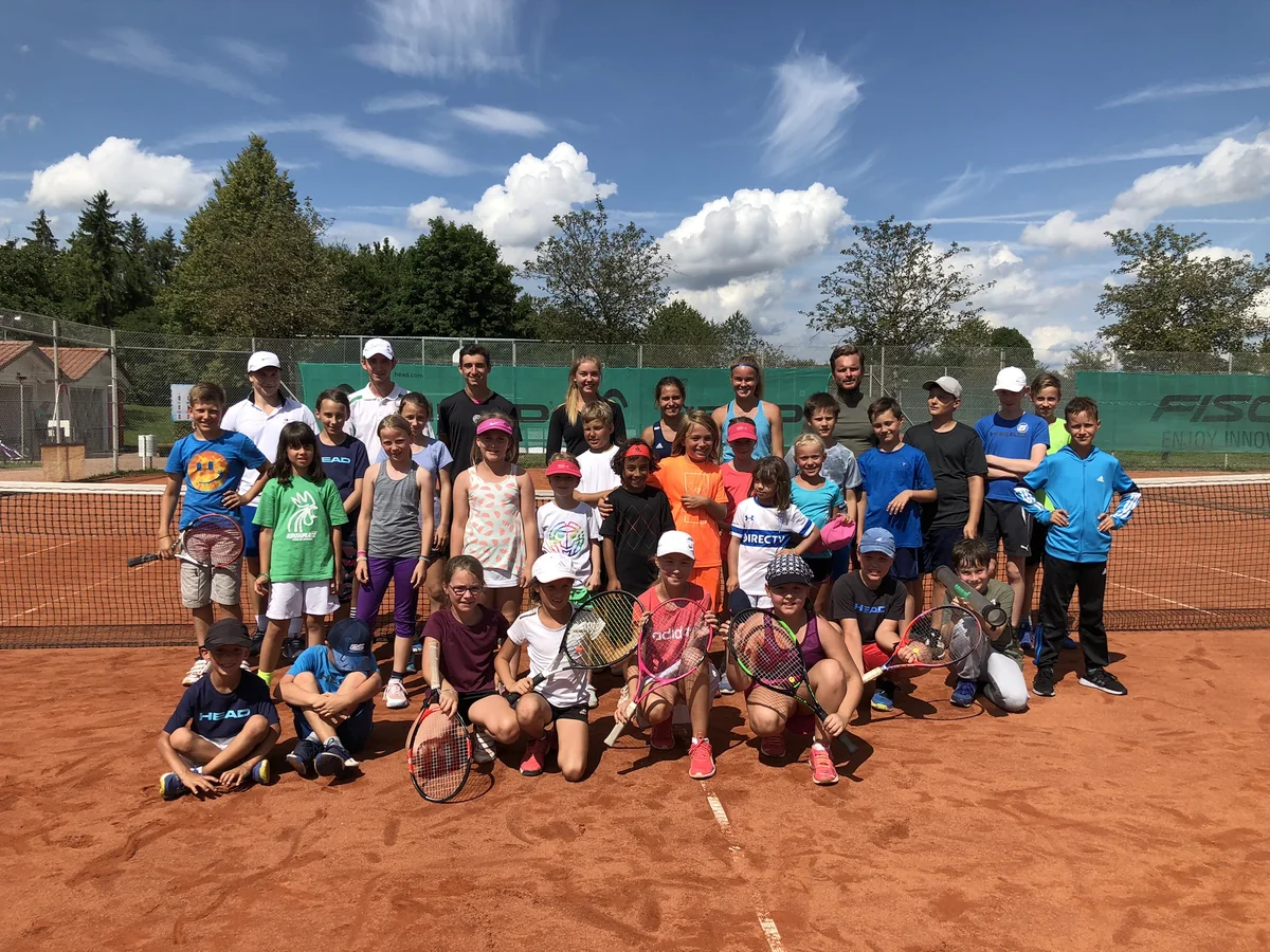 Große Gruppe Kinder und Jugendlicher mit Trainerteam auf einem Sandplatz vor sommerlichem Himmel, locker aufgestellt