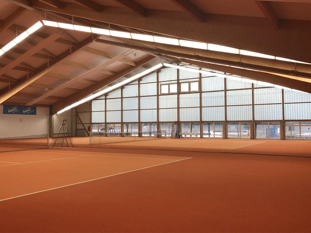 Blick in die TCI-Tennishalle: Holzdachstuhl, helle Glasfront und zwei Sandplätze nebeneinander.