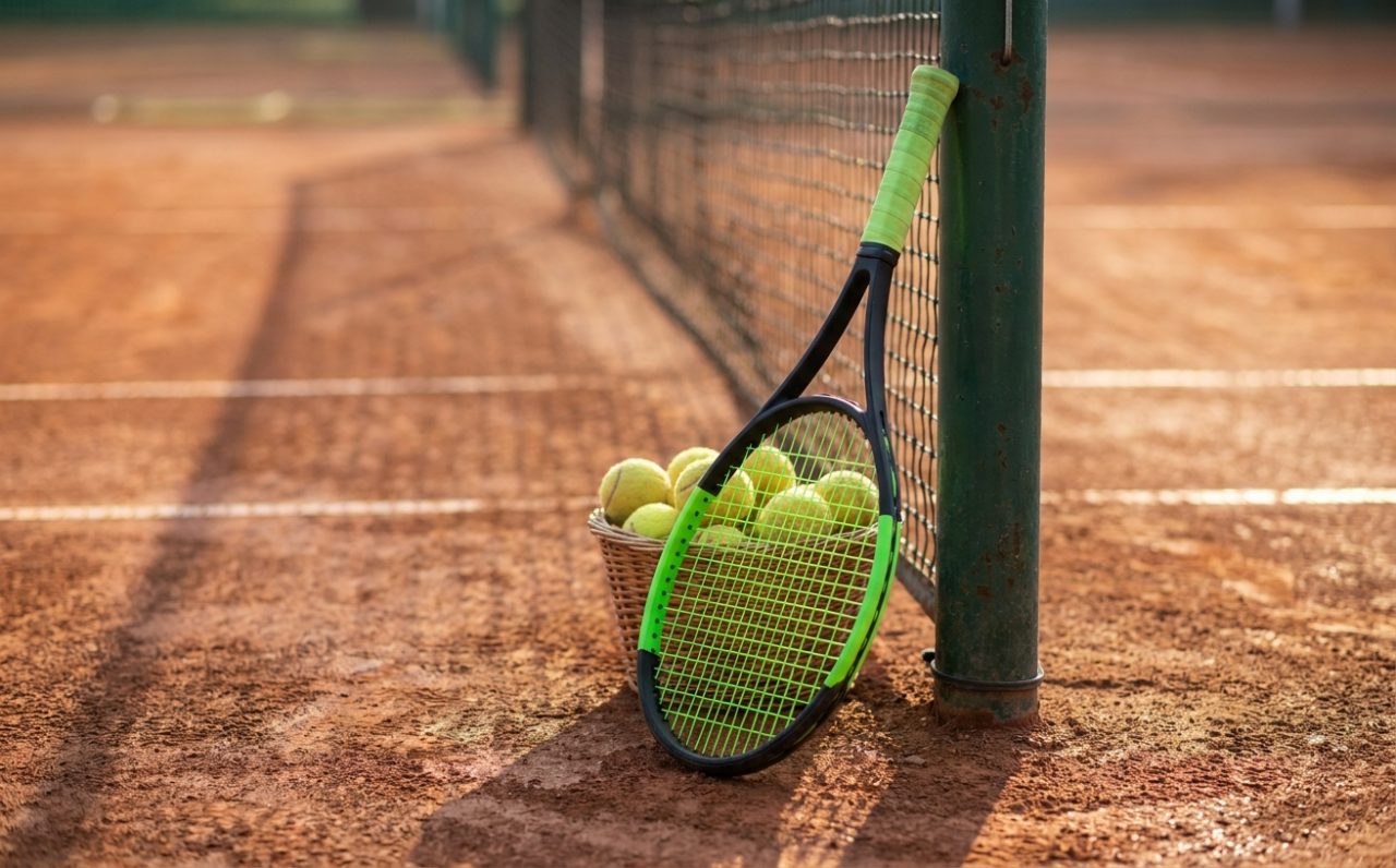Moderner Tennisschläger und ein Ballkorb am Netzpfosten auf ruhigem Sandplatz im Morgenlicht