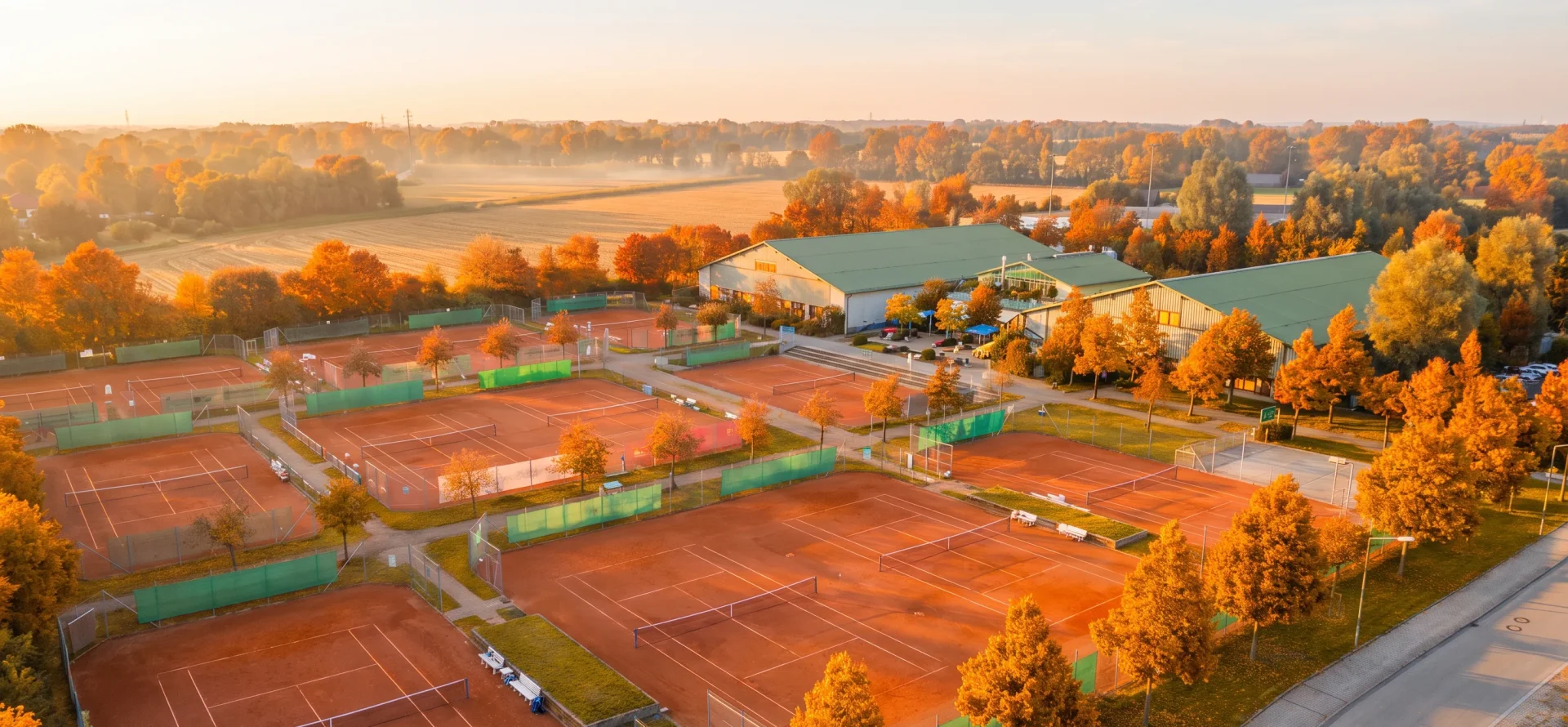 Luftaufnahme der TCI-Anlage im Herbst – Sandplätze und Hallen zwischen Bäumen in leuchtendem Orange und Gold, dahinter abgeerntete Felder im Morgendunst.